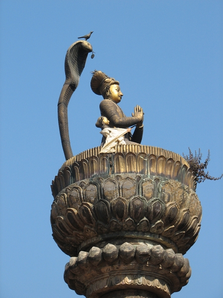 IMG_8466.JPG - King Yoganarendra Malla's statue - Outside the Hari Shankar Temple is a tall column topped by a figure of King Yoganarendra Malla and his queens. The golden figure of the kneeling king atop a lotus bud and protected by the hood of a cobra has been facing towards his palace since 1700.  On the cobra's head is a bird figure; legend has it that as long as the bird remains there the king may still return to his palace. A door and window of the palace are always ket open and a hookah is kept ready for the king should he return.